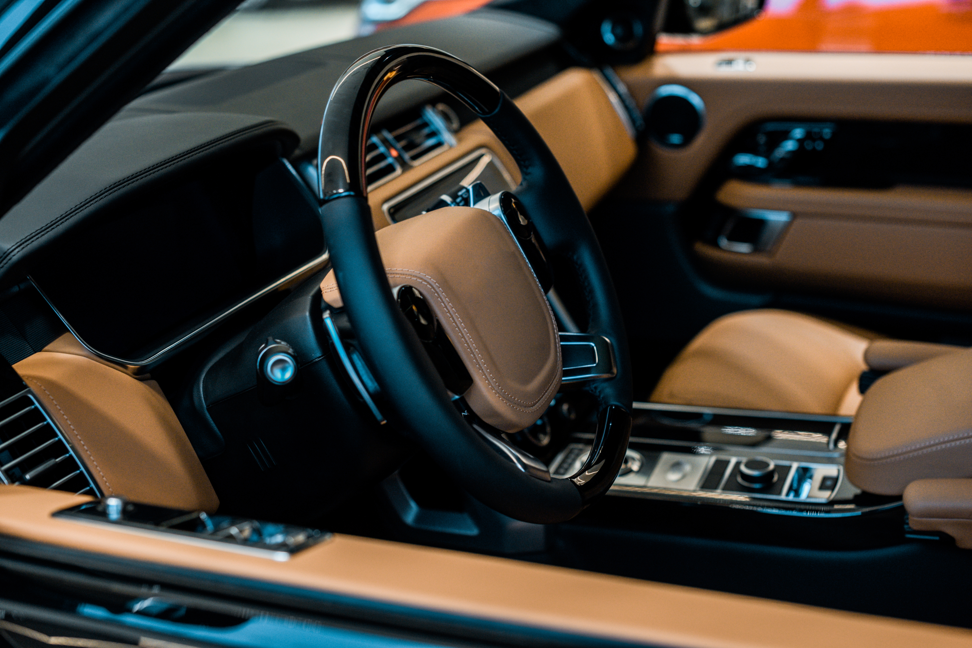 Nahaufnahme eines modernen Luxusauto-Innenraums mit schwarzem Lenkrad und Armaturenbrett sowie hellbraunen Ledersitzen und -verkleidungen, die ein elegantes Design und hochwertige Materialien präsentieren.
