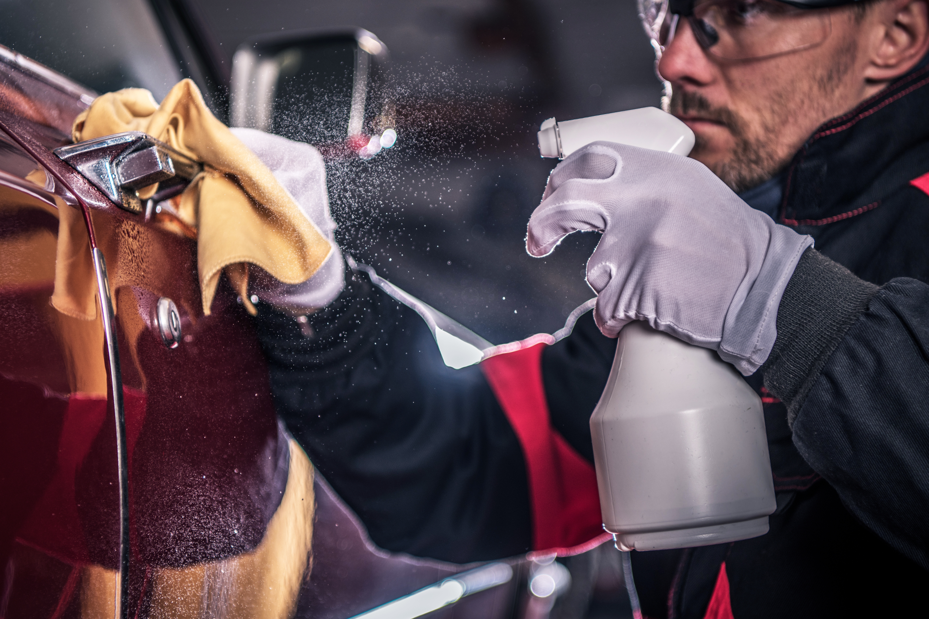Eine Person mit Handschuhen und Schutzbrille sprüht eine Reinigungslösung auf ein rotes Auto und wischt es mit einem gelben Tuch ab, um das Äußere des Fahrzeugs zu säubern.